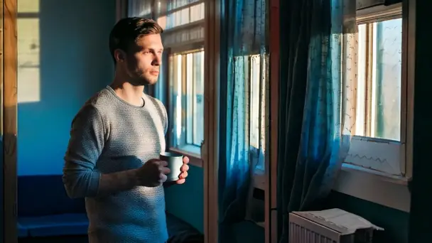 A man standing at a living room window in the morning holding a mug in both hands, looking outside