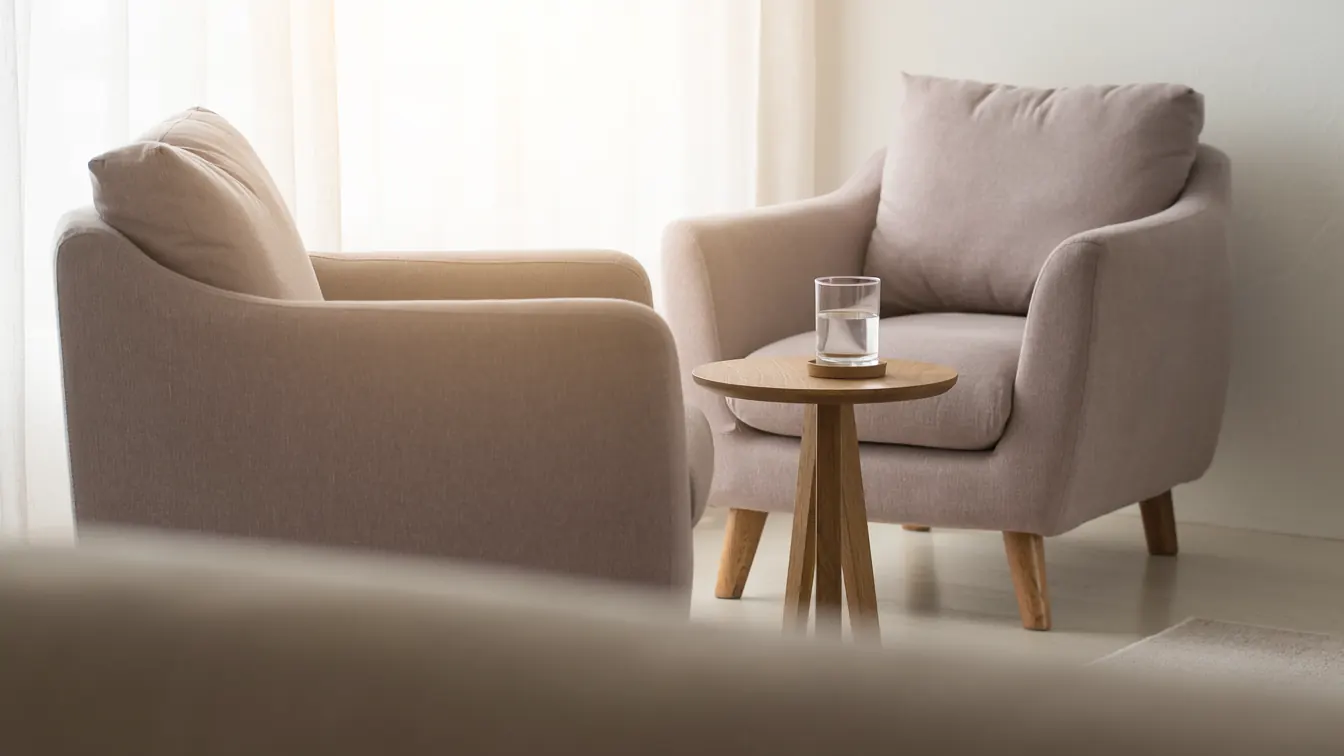 Two pale armchairs facing each other beside a bright window, a glass of water on a small table between them