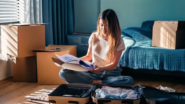A young woman sitting on a bedroom floor beside packing boxes and an open suitcase, reading a page from a folder
